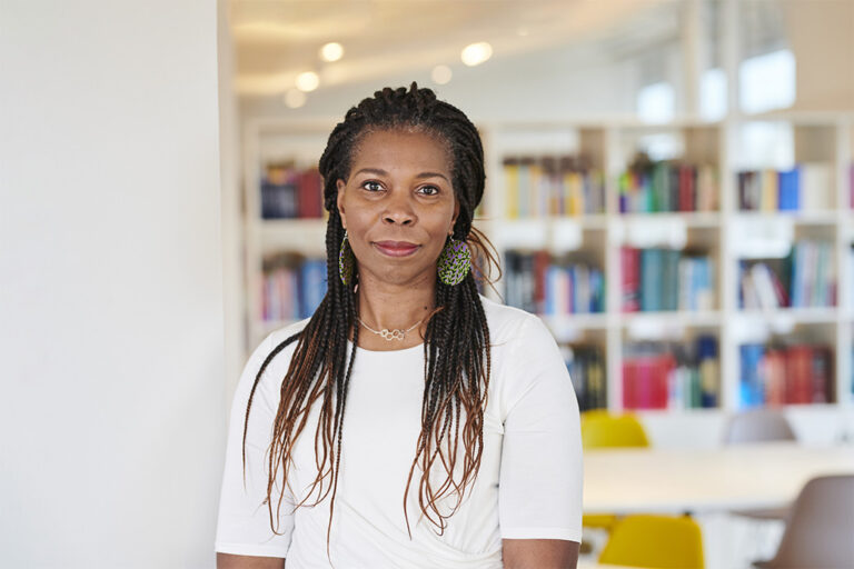 Tase Oputu stands in front of a bookcase, wearing a white top