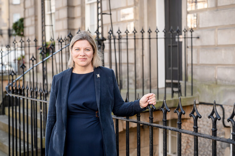 Laura Wilson stands by a rail, wearing a blue dress and blazer
