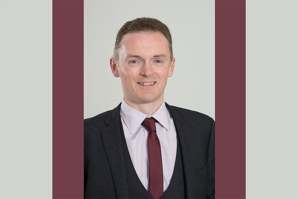 Ryan smiles at the camera, wearing a black blazer and plum coloured tie