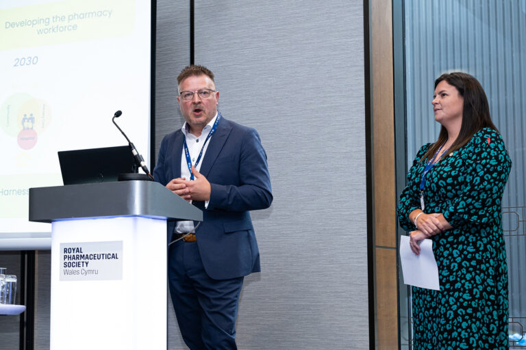 Jonathan Simms is speaking at the podium and Elen Jones watches on