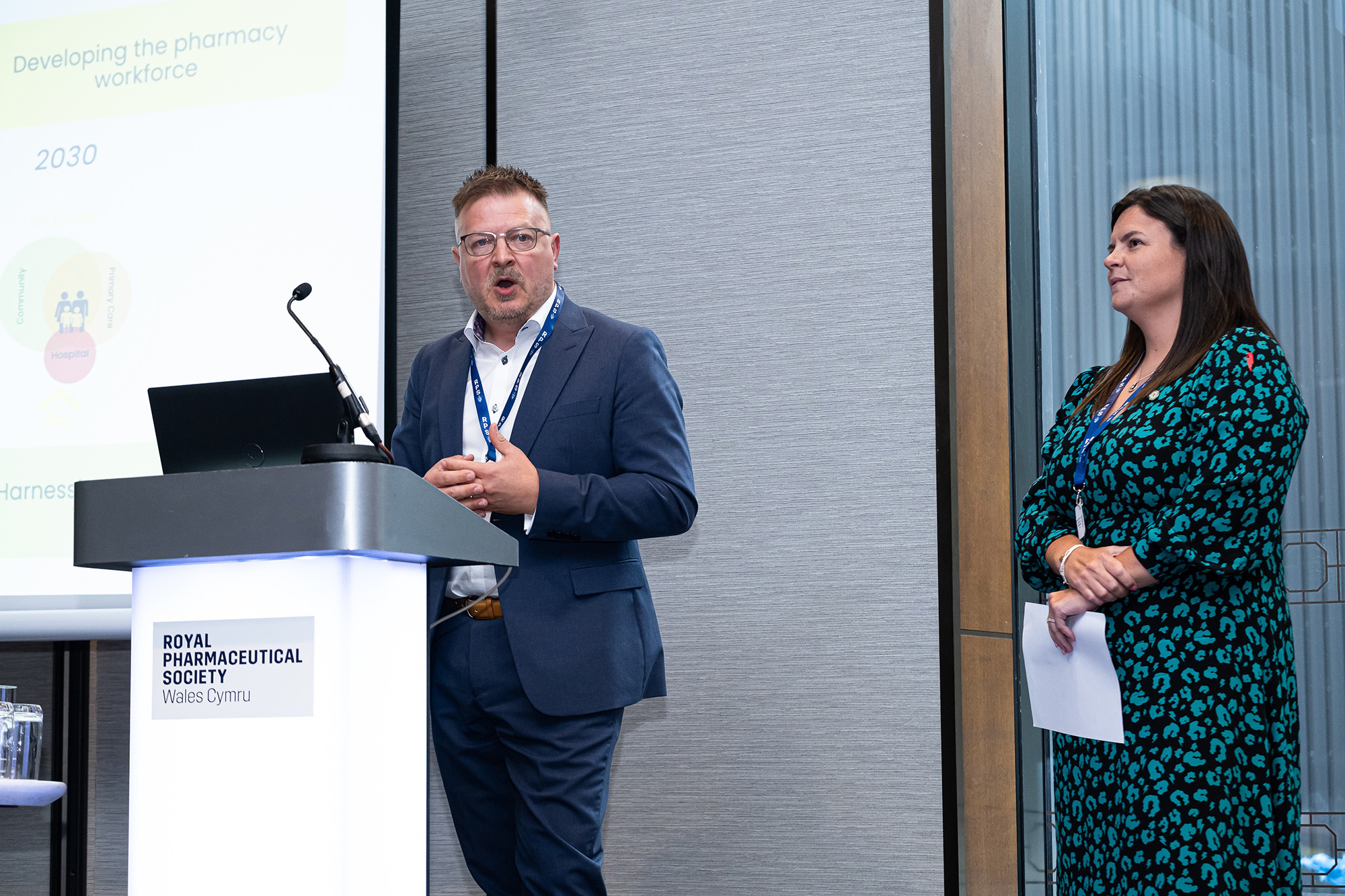 Jonathan Simms is speaking at the podium and Elen Jones watches on