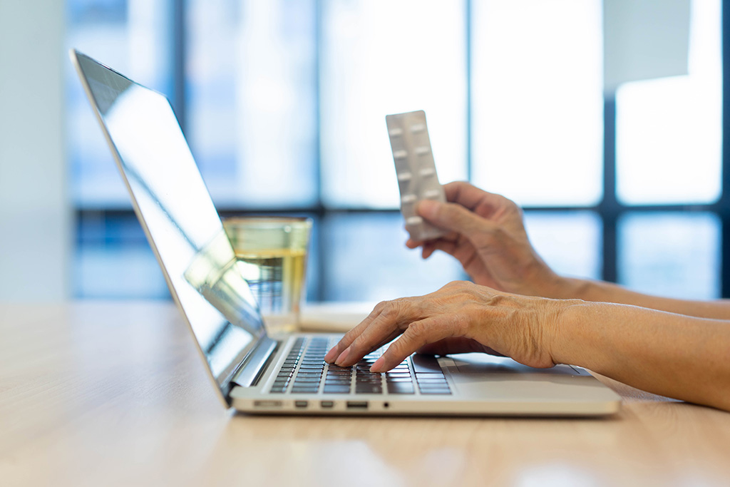 Laptop open on a bench, side view. Person types and holds pill packet up