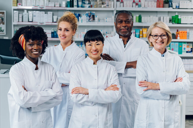 A multicultural team of five professional pharmacists stand together in a pharmacy