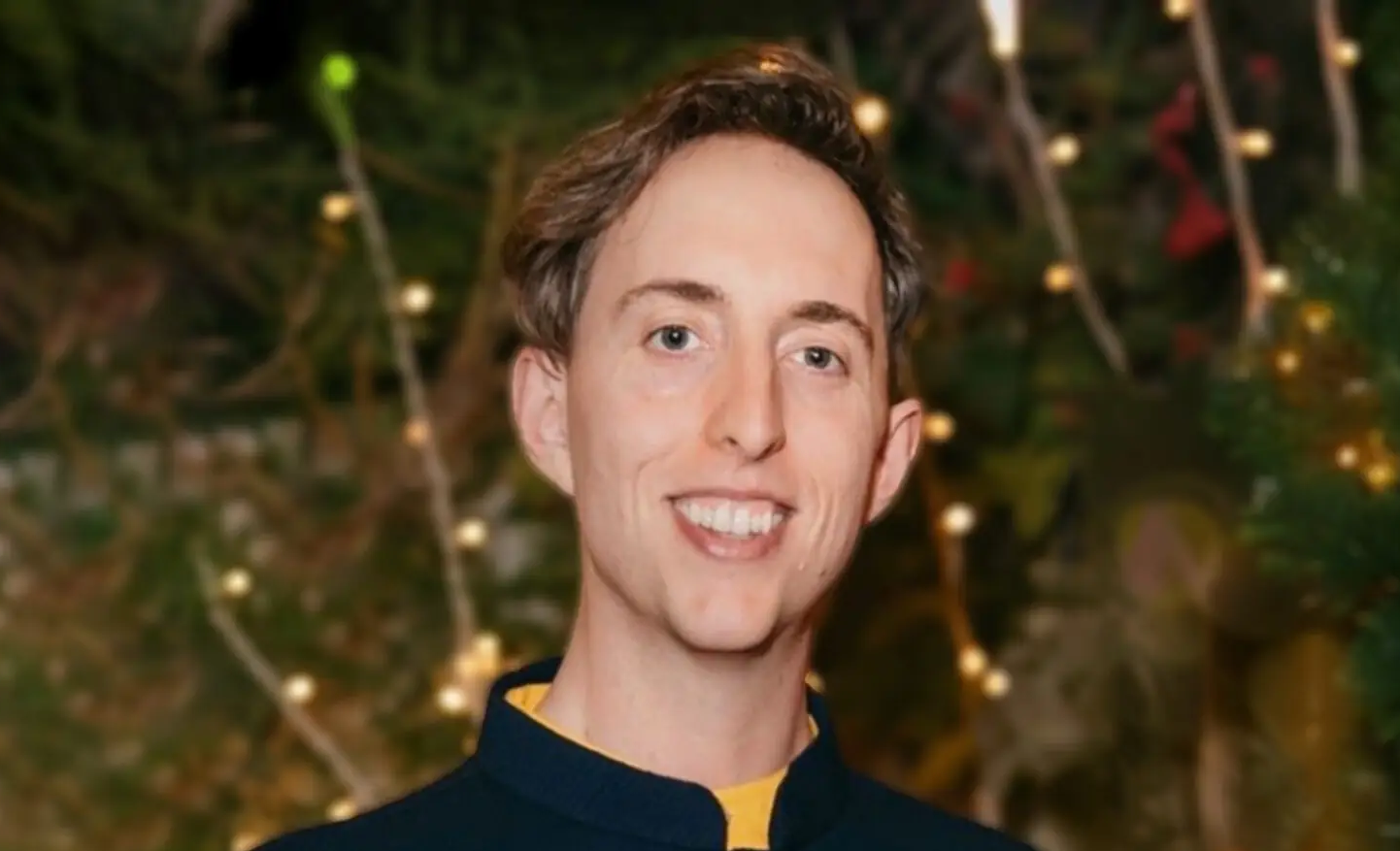 Adam smiles in front of a dark background with trees and lights