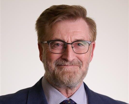 A portrait of a man in a blue suit and tie smiling with black glasses with a grey beard and short red hair.
