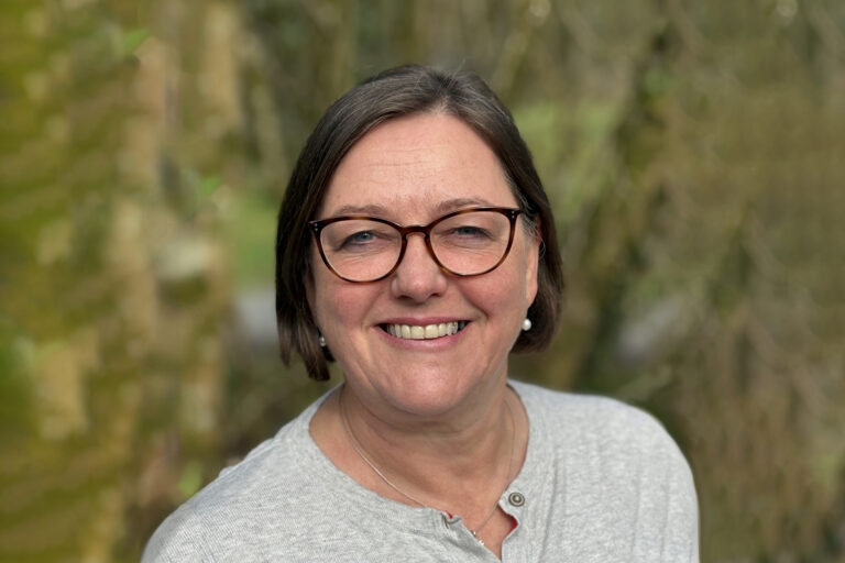 Emily smiles wearing glasses and a grey top in front of a blurred green nature background