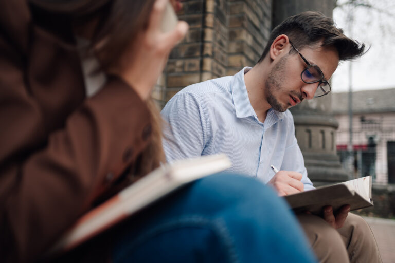 Two college students sitting outdoors on campus, focused on writing in their notebooks