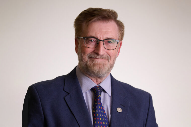 Joe smiles wearing glasses and a suit in front of a neutral background