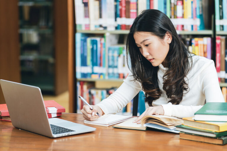 Asian female student study in a library and using laptop. Bookshelves behind