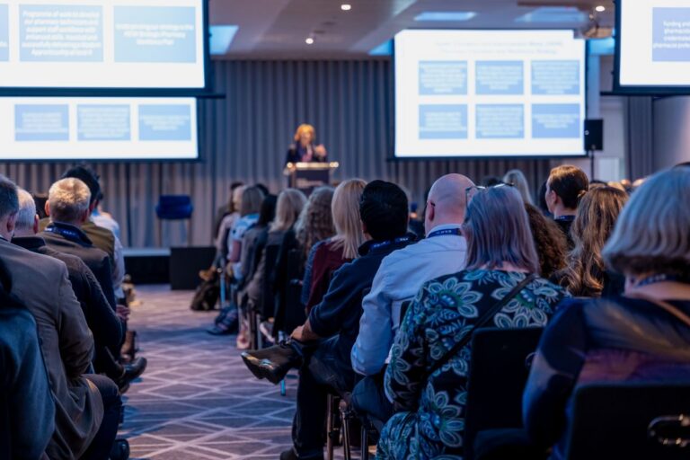 A speaker at RCPharm's annual conference 2024 surrounded by large white screens with blue boxes while members of the audience watch in the conference hall