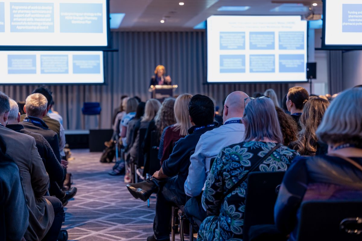 A speaker at RCPharm's annual conference 2024 surrounded by large white screens with blue boxes while members of the audience watch in the conference hall