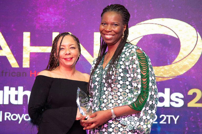 Tase and Professor Patricia Miller OBE - Patron of the Black Healthcare Awards, smile in front of purple screen holding a glass award