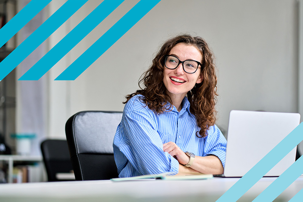 A young woman wearing glasses sat at a desk with her laptop open smiles and looks to the left. Dark and light teal stripes frame the image (Source: insta_photos / Shutterstock.com)