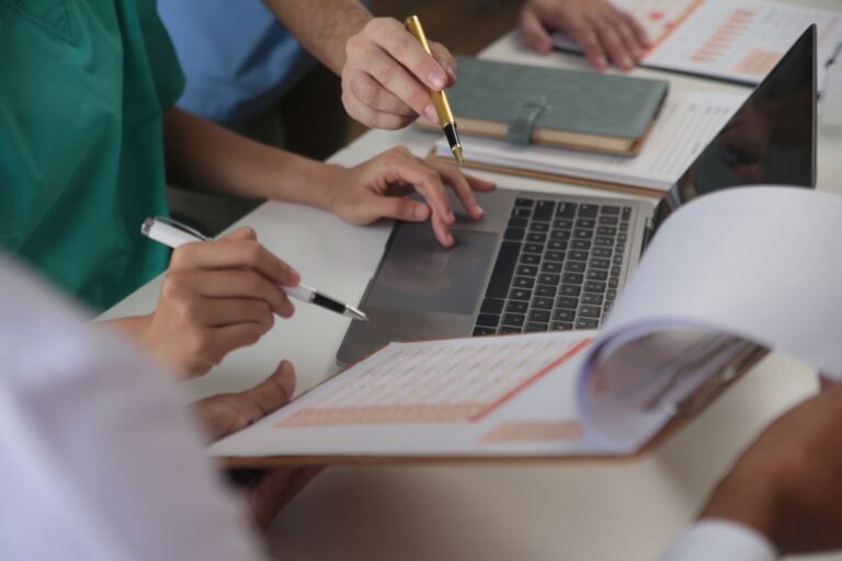 Medical team meeting around table in modern hospital, close up of hands holding pens above a laptop keyboard