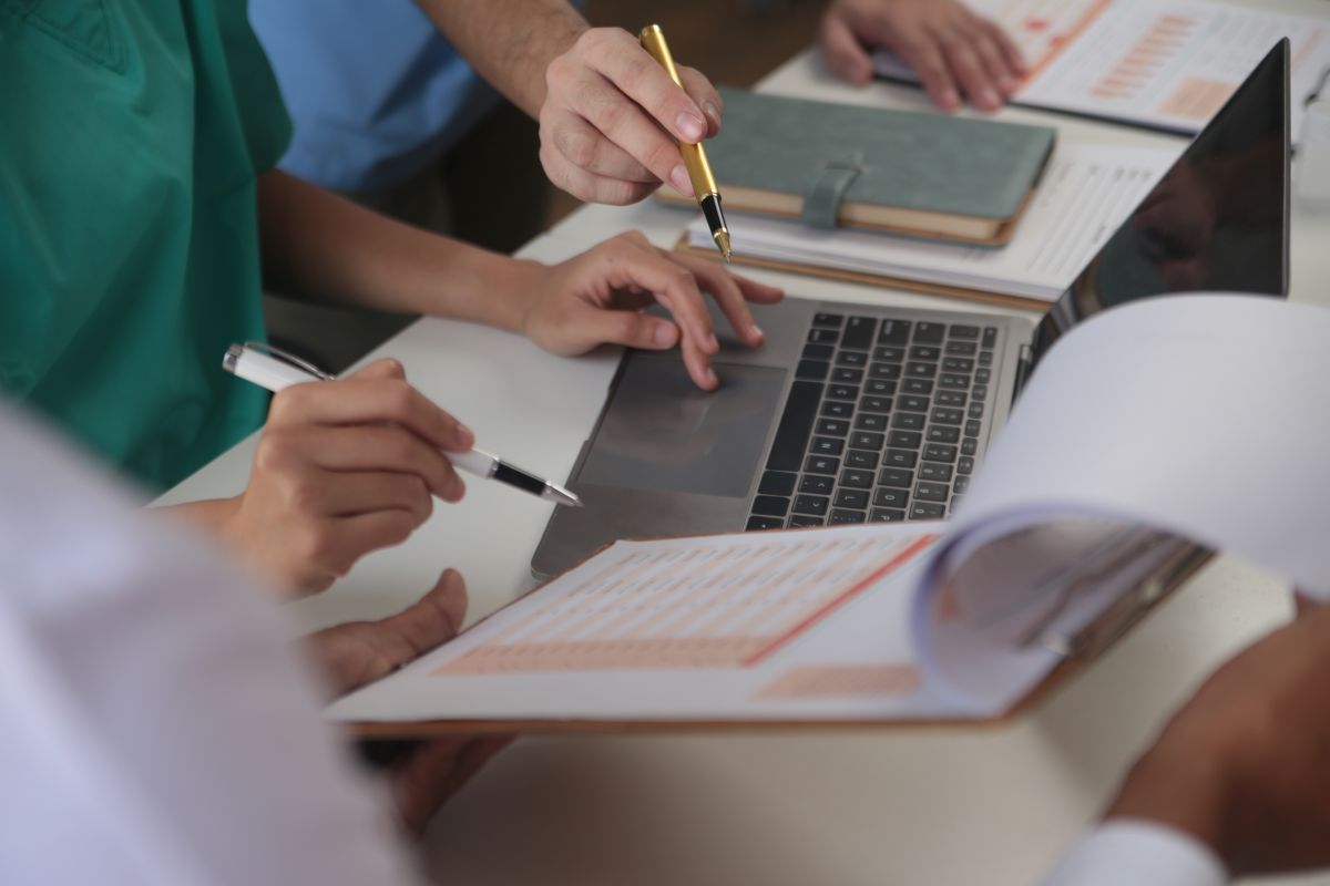 Medical team meeting around table in modern hospital, close up of hands holding pens above a laptop keyboard