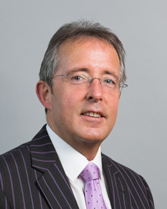 Head and shoulders image of Dr Chris Martin OBE, Chair of the ‘Pharmacy: Delivering a Healthier Wales’ delivery board, wearing a striped suit and lilac tie