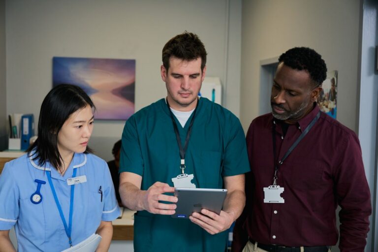 A nurse and two other healthcare professionals stand together in a hospital corridor, reviewing information on a tablet.