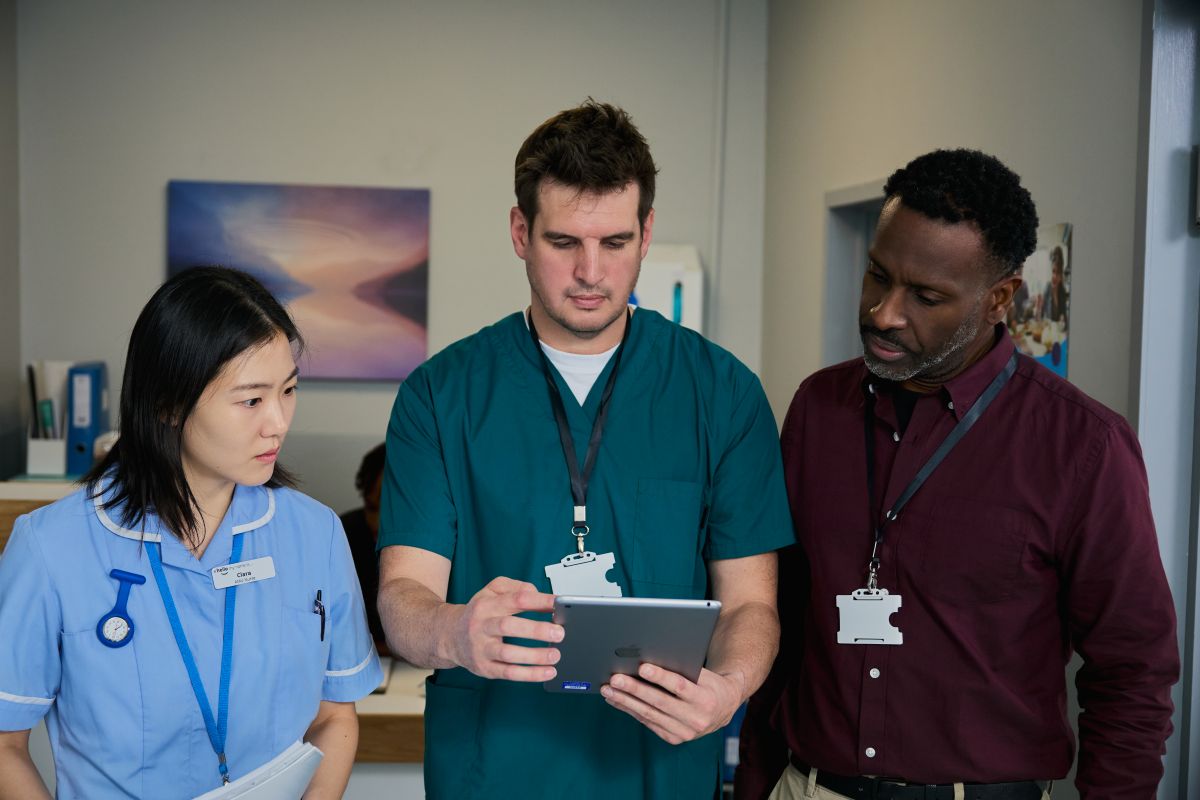 A nurse and two other healthcare professionals stand together in a hospital corridor, reviewing information on a tablet.