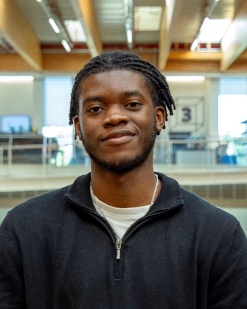Head and shoulders image of Jaden Sanni, member of the ‘Pharmacy: Delivering a Healthier Wales’ delivery board, wearing a black top over a white tee shirt 