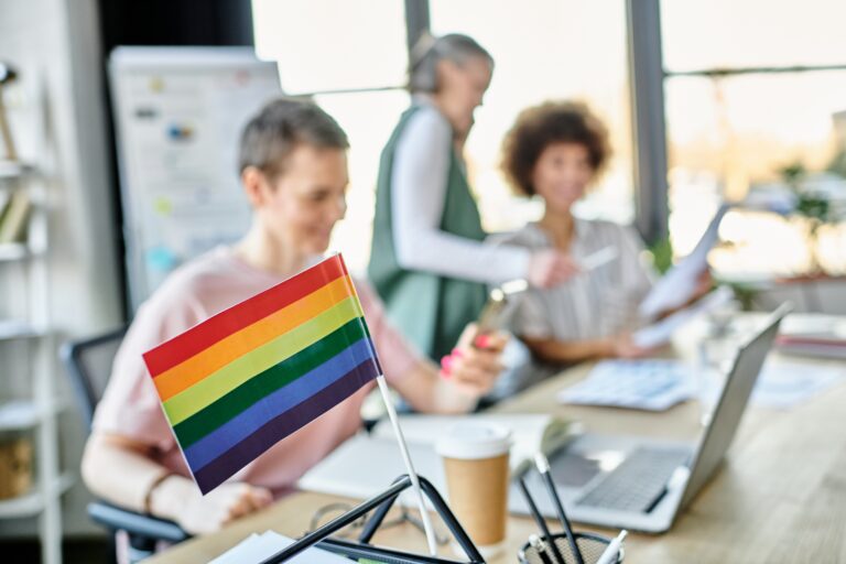 Focus on rainbow flag displayed on office table in front of blurred diverse businesswomen.