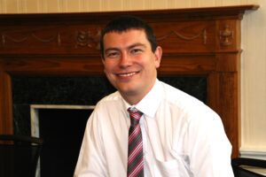 Image of Matt Barclay FRCPharm, Chief Executive Officer, Community Pharmacy Scotland, wearing a white shirt and striped tie in front of a fireplace