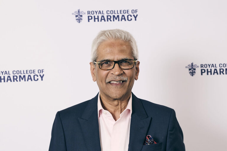 Mahendra Patel smiles in front of a white banner with navy RCPharm logos. He wears a light pink shirt with a dark blazer and a red patterned pocket square.