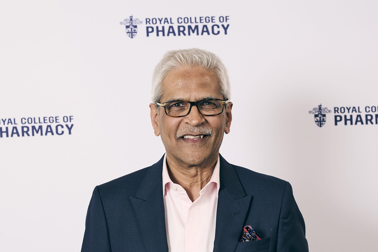 Mahendra Patel smiles in front of a white banner with navy RCPharm logos. He wears a light pink shirt with a dark blazer and a red patterned pocket square.