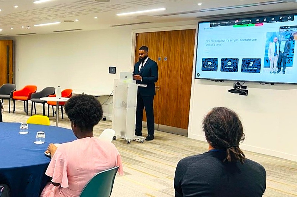 Samson Akere standing at a white lectern with a large presentation screen to his left. He is addressing a room of people sitting at round tables.